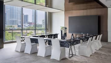 Una sala de conferencias moderna con una gran mesa, sillas blancas, una pantalla de proyector y ventanas amplias que ofrecen vistas a la ciudad.