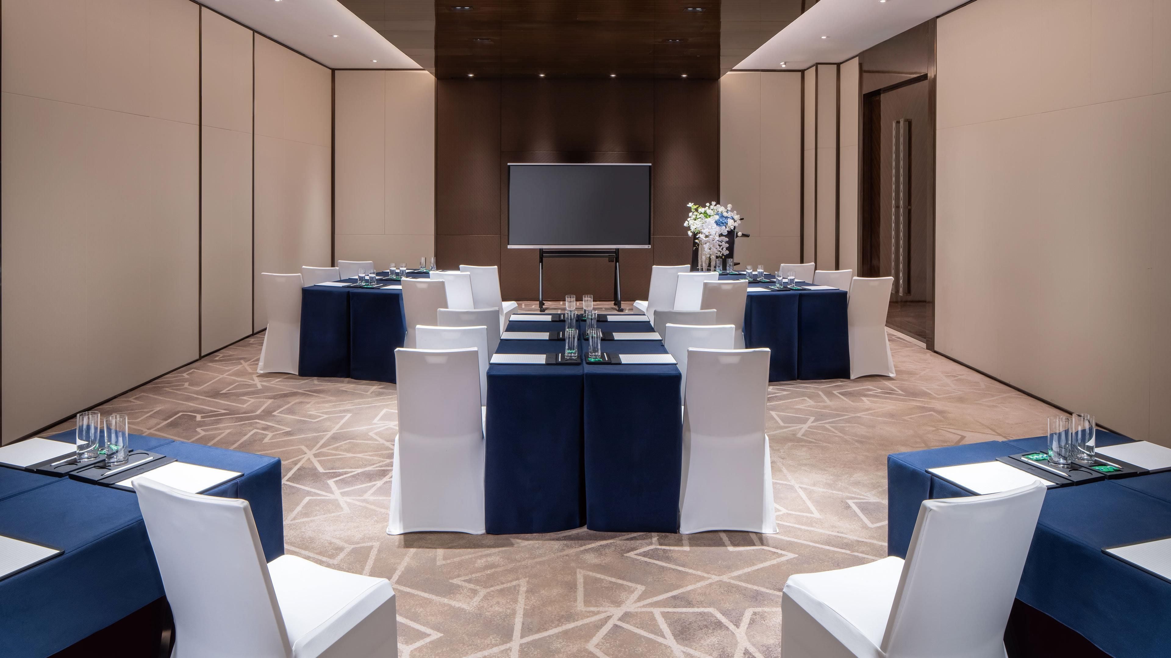 Un salón de conferencias con una pantalla grande, varias mesas con sillas y una alfombra estampada.