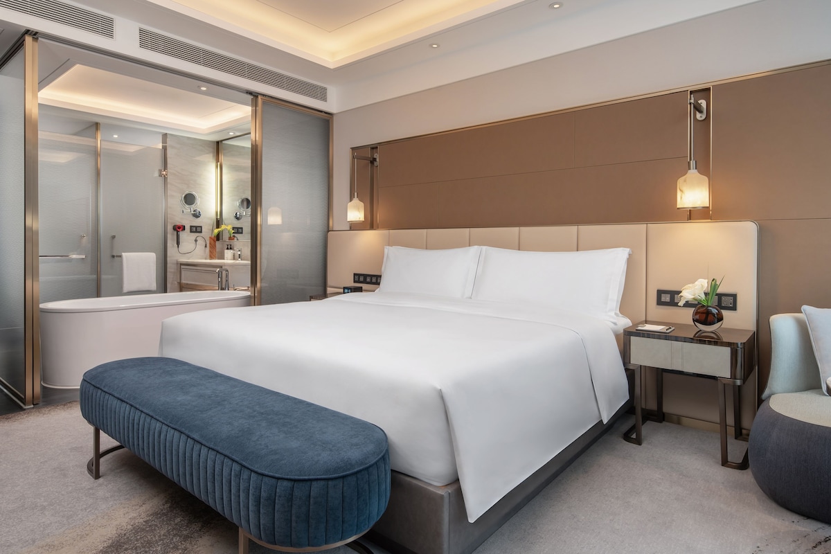 Une chambre d’hôtel moderne dotée d’un grand lit, d’une salle de bain séparée, d’un coin salon et d’une table de chevet avec une lampe.