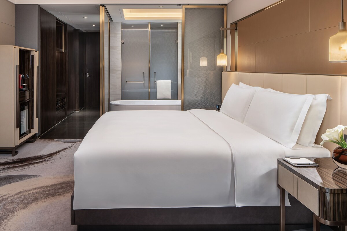 A modern hotel room with a large bed, a walk-in closet, a shower area, and a bedside table with a lamp.