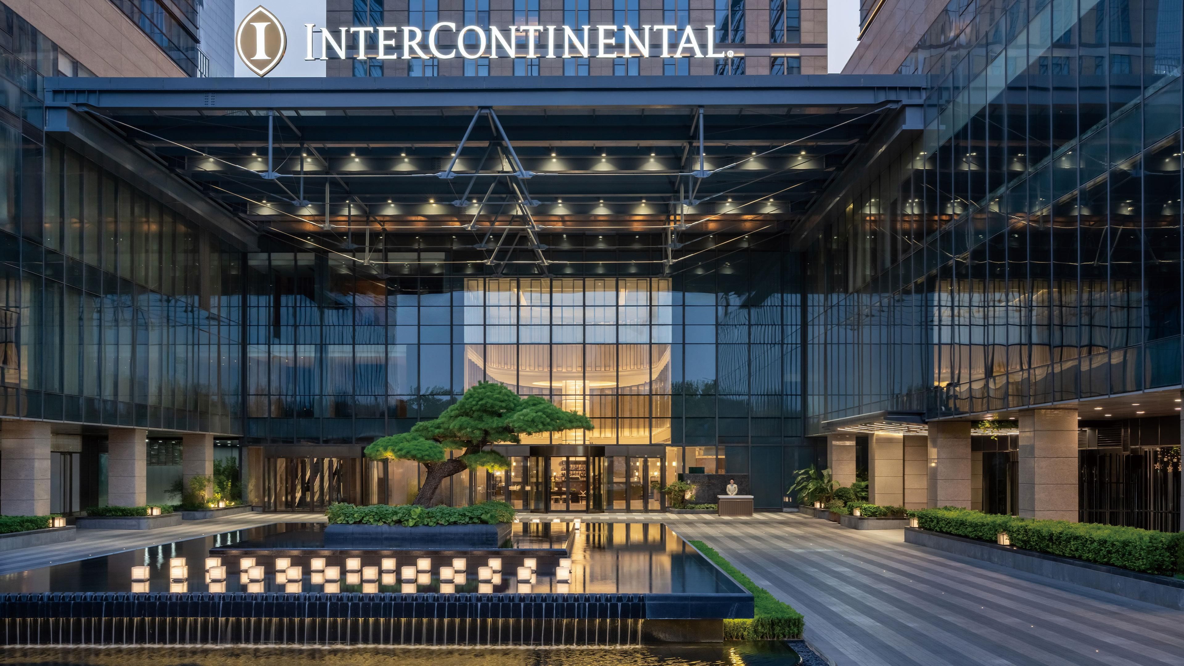 The Intercontinental hotel with a modern glass facade, a central water feature, and a well-lit entrance.