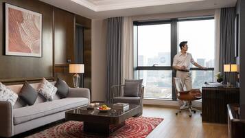 Habitación de hotel moderna con sofá, escritorio, silla, mesa baja con frutero y ventana grande con vista a la ciudad.