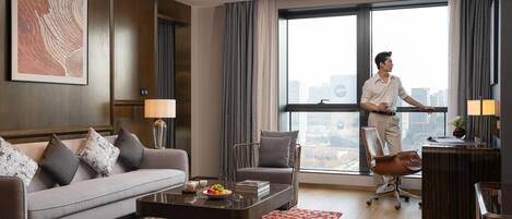 A modern hotel room with a sofa, a desk, a chair, a coffee table with a fruit bowl, and a large window with a city view.
