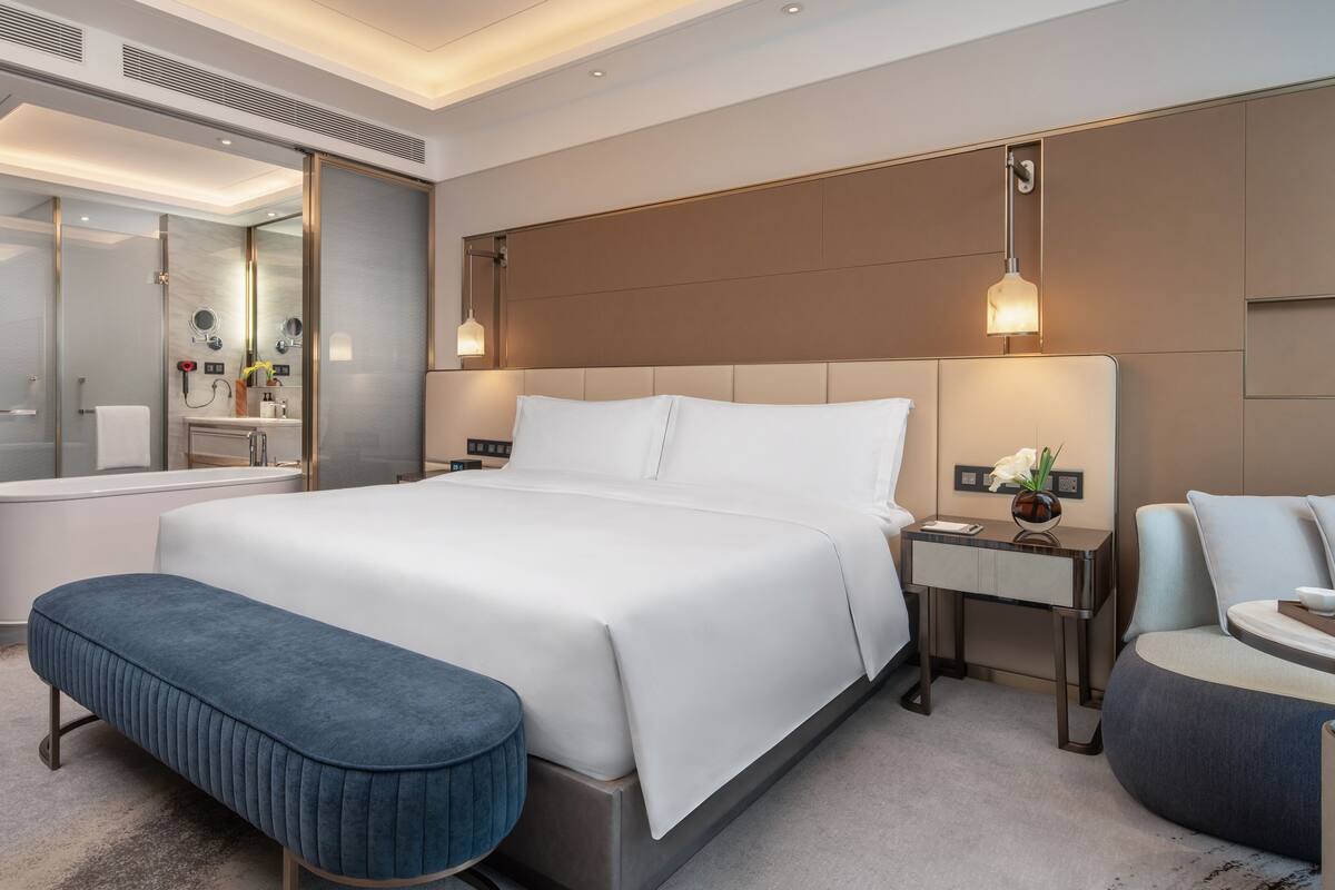 Una habitación de hotel moderna con una cama grande, baño independiente, zona de estar y una mesita de noche con lámpara.