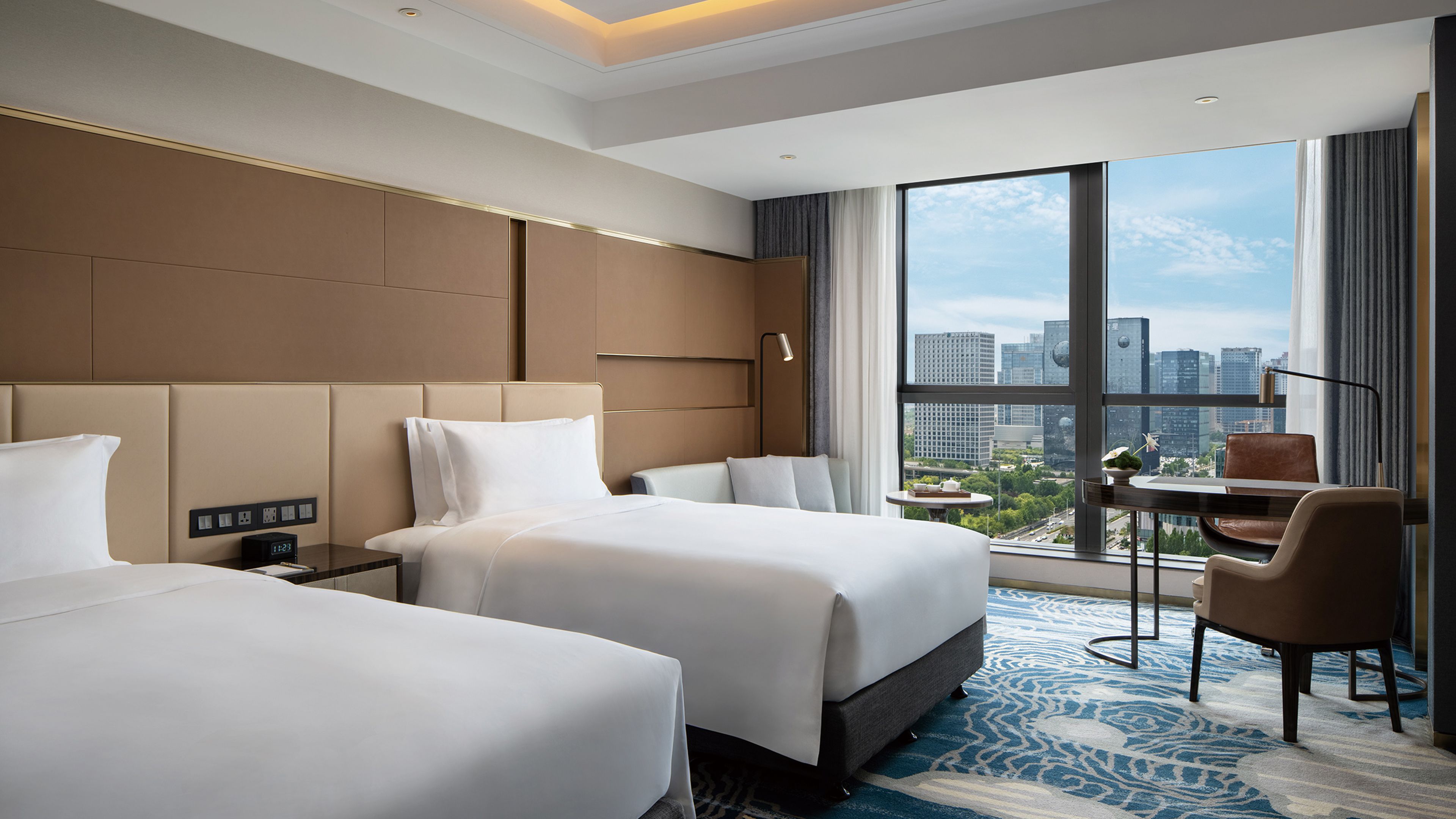 A hotel room with two beds, a desk, and a chair. There is a large window with a city view.