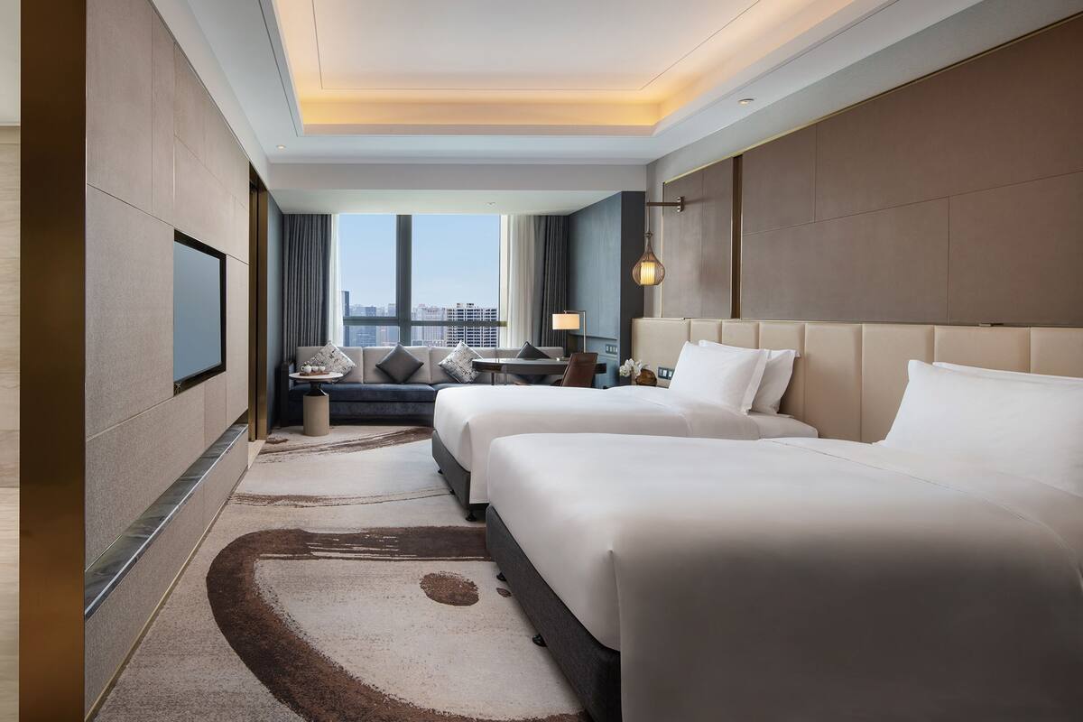 Habitación de hotel con dos camas, un sofá, un escritorio y un amplio ventanal con vista a la ciudad.