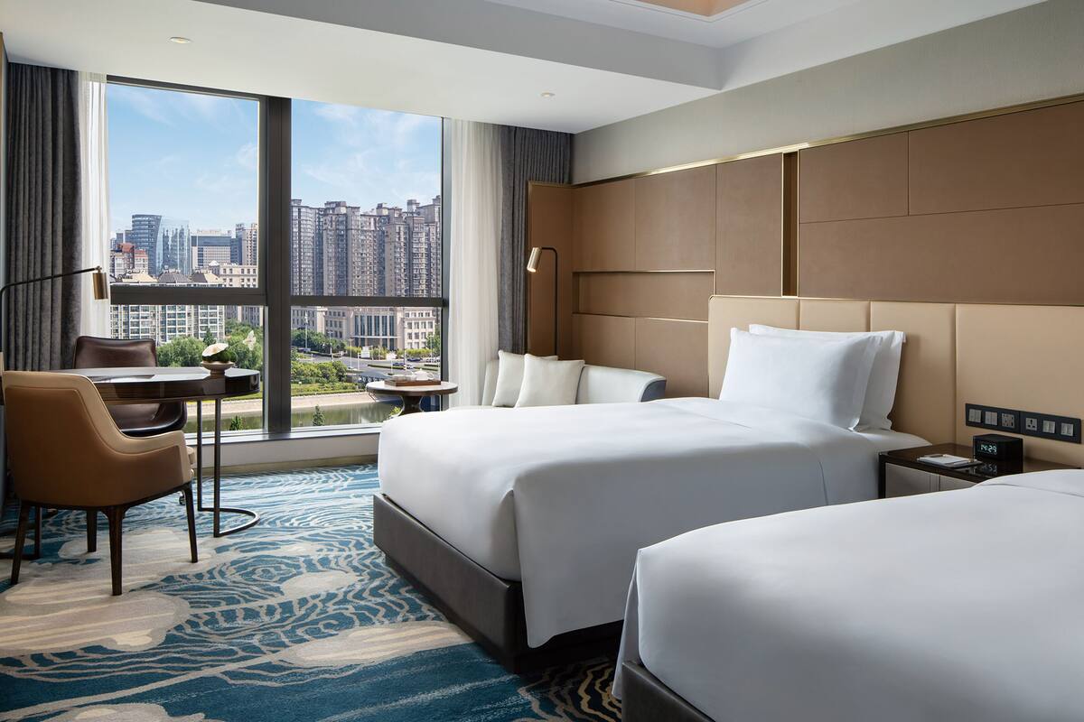 Habitación de hotel con dos camas, un escritorio, una silla, una mesita de noche y un ventanal grande con vistas a la ciudad.