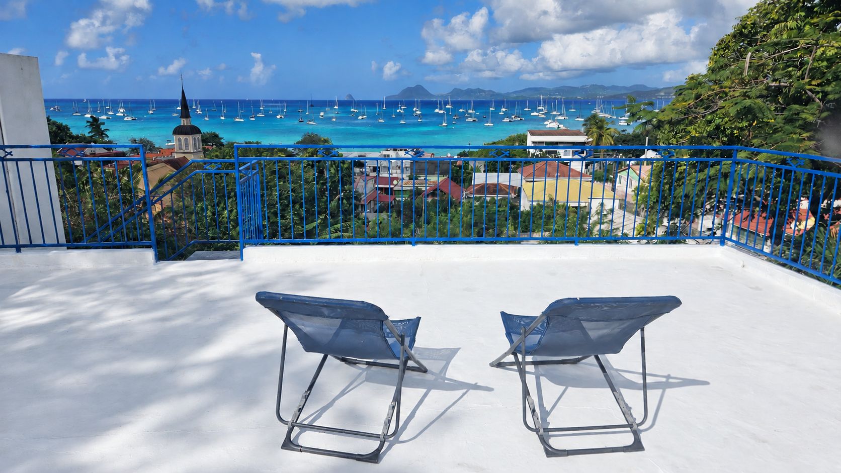 A balcony with two blue chairs, a clear view of the sea with boats, and a church in the background.