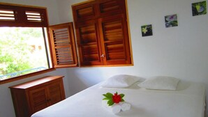 A hotel room with a bed, a window with wooden shutters, a small wooden cabinet, and a wall with three small flower paintings. - Villa with sea view in the heart of Sainte-Anne (Martinique)