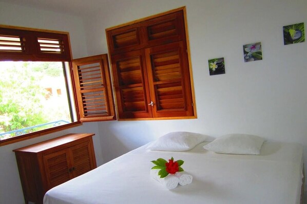 A hotel room with a bed, a window with wooden shutters, a small wooden cabinet, and a wall with three small flower paintings. - Villa with sea view in the heart of Sainte-Anne (Martinique)