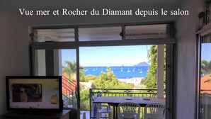 A room with a view of the sea and a mountain, a balcony with a table and chairs, and a television on a cabinet. - Villa with sea view in the heart of Sainte-Anne (Martinique)