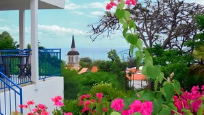 A balcony with a blue railing overlooking a coastal area with a church and lush greenery. - Villa with sea view in the heart of Sainte-Anne (Martinique)
