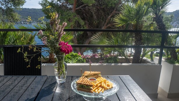 A table with a glass dish of food, a vase with flowers, and a view of the sea and trees. - Hotel Issa - Double Room Park View 26 (Vis)