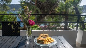 Una mesa con un recipiente de vidrio con comida, un florero con flores y vista al mar y a los árboles.