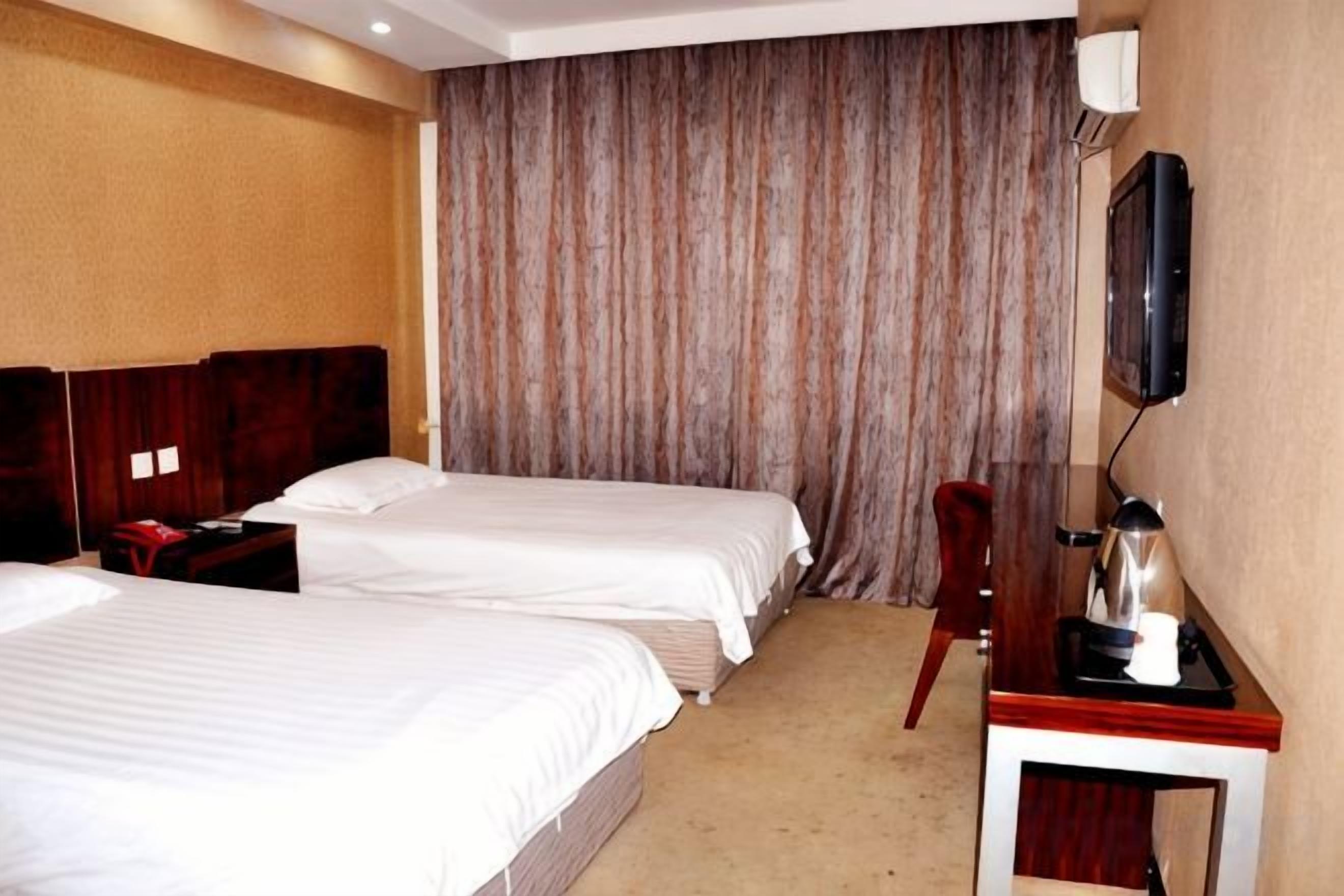A hotel room with two beds, a desk with a kettle, and a large window with curtains.