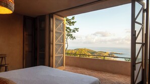 A bedroom with a large bed, a wooden chair, and a balcony with a view of the sea and hills. - Monte LaLaLa - Eco-Lodge à Mermejita - Vue imprenable depuis la piscine! (Oax.)
