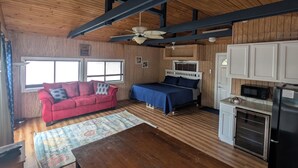Ein gemütliches Blockhaushaus-Interieur mit Holzbalkendecke, einem roten Sofa, einem Bett mit blauer Tagesdecke, einer Küchenzeile mit Mikrowelle und einem Esstisch.