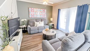 Een moderne woonkamer met een grijze bank, een salontafel en een televisie. Er is een plafondventilator, een schilderij aan de muur en een raam met gordijnen.