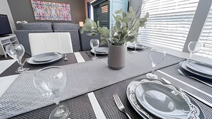 Een eettafel gedekt met borden, glazen en bestek, een vaas met groene planten en een moderne bank op de achtergrond.