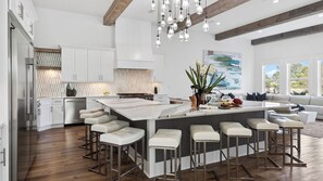 Une cuisine moderne avec des meubles blancs, un grand îlot central doté de tabourets, et un coin repas comprenant un canapé et de grandes fenêtres.