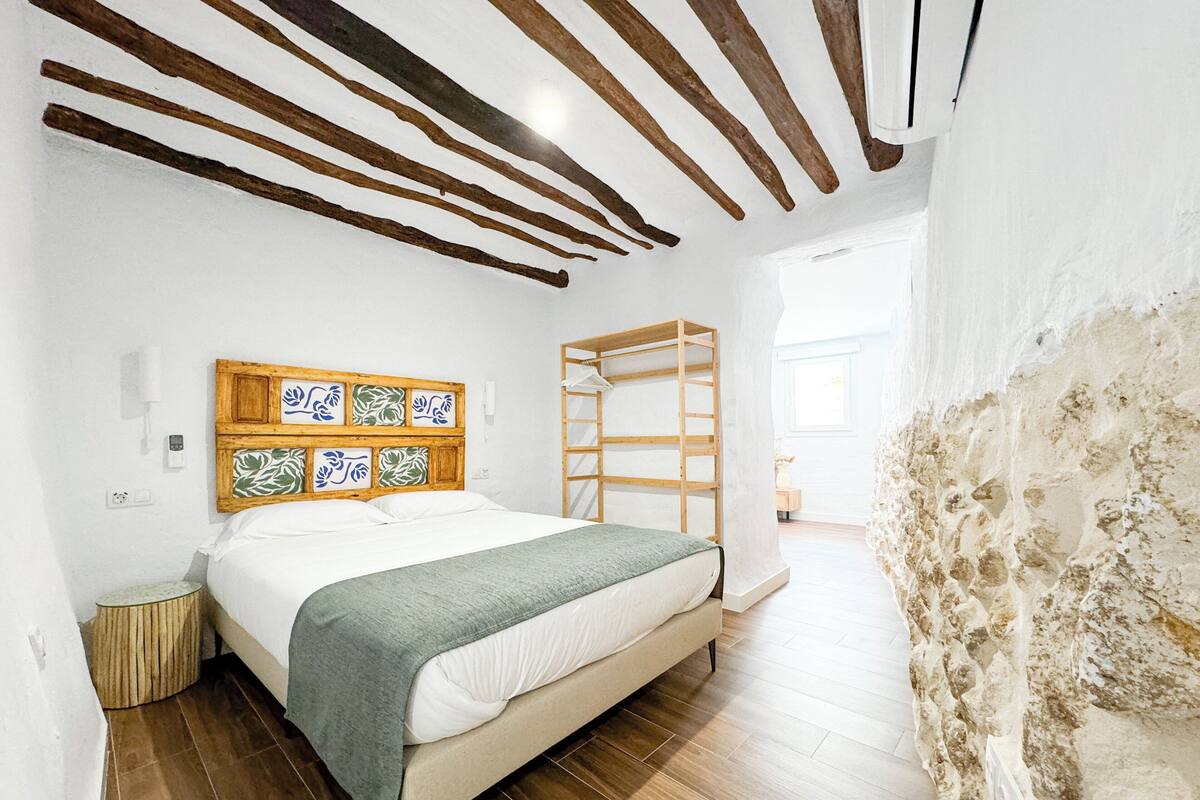 A bedroom with a wooden headboard, a bed with a green blanket, a wooden ladder, and a stone wall.
