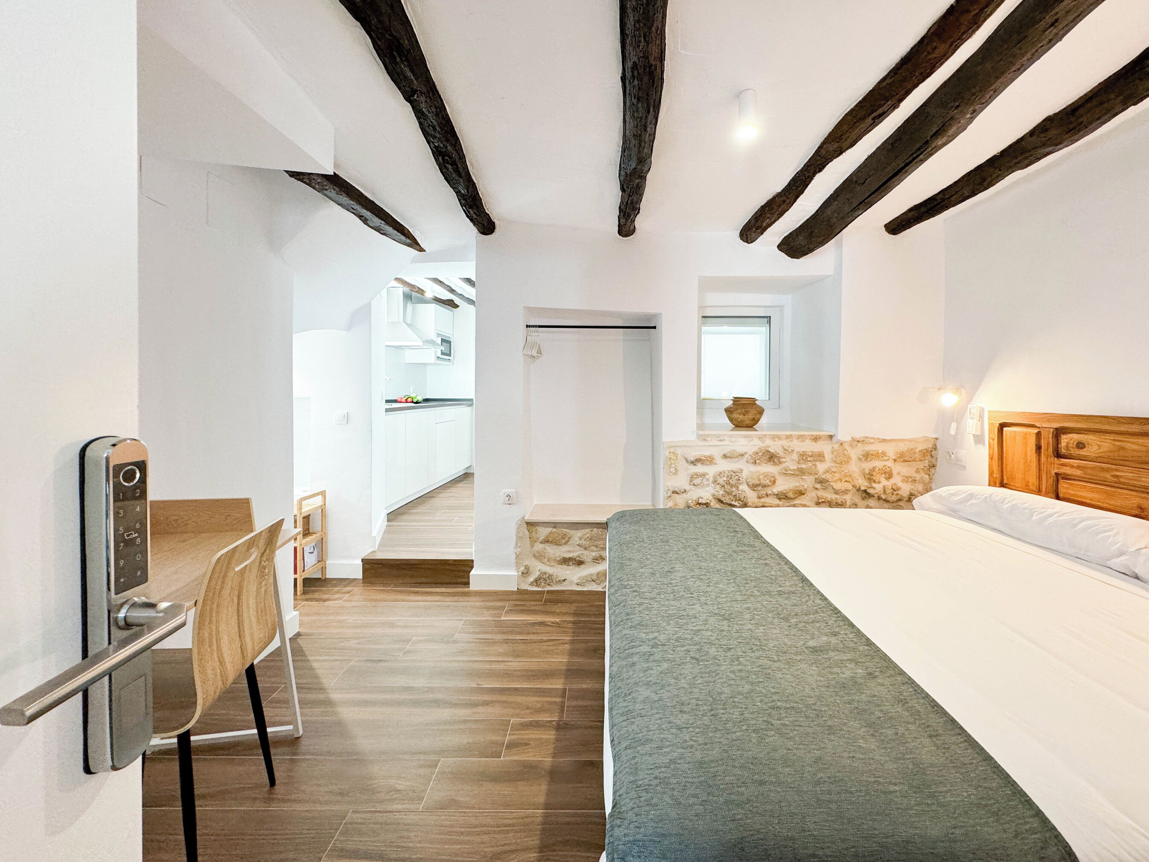 A bedroom with a bed, a desk, a chair, and a view of a kitchen and bathroom.