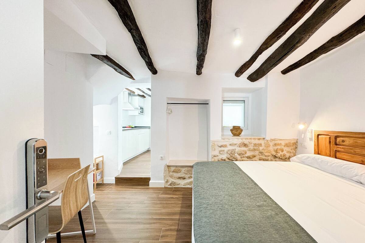 A bedroom with a bed, a desk, a chair, and a view of a kitchen and bathroom.