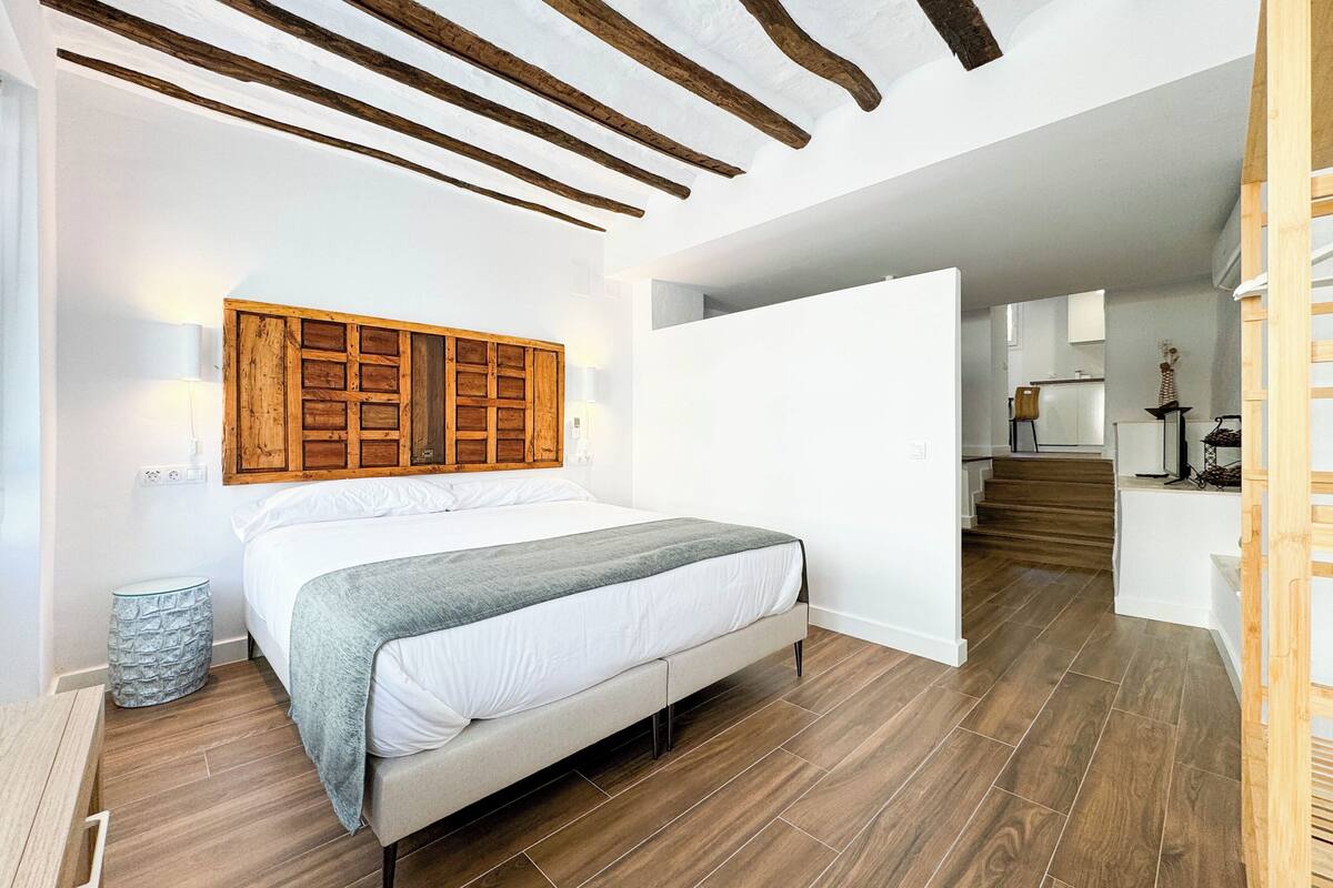 A bedroom with a wooden headboard, a bed with white linens, a grey throw, and a wooden floor.
