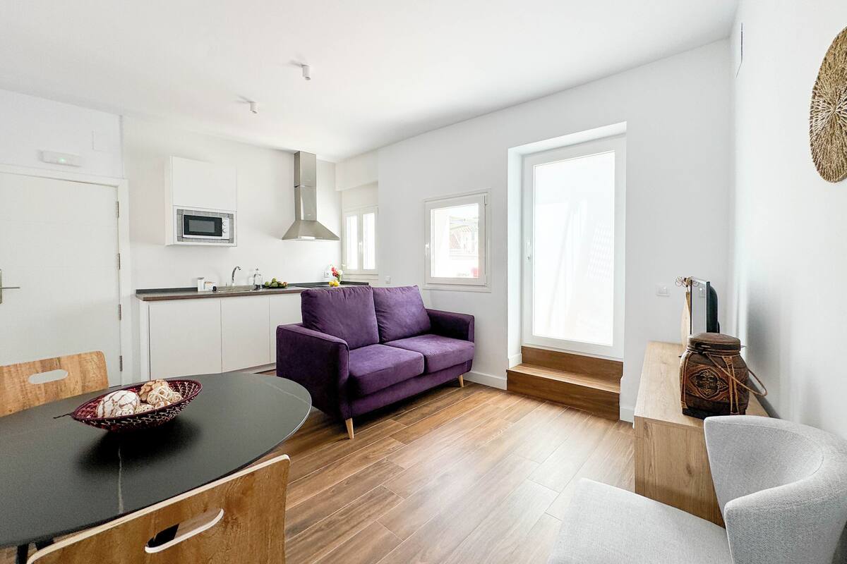 A modern apartment with a kitchen, a purple sofa, a dining table, and a large window.