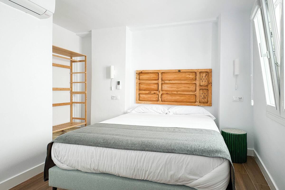A modern bedroom with a wooden headboard, a bed with white and grey bedding, a wooden ladder, and a green stool.