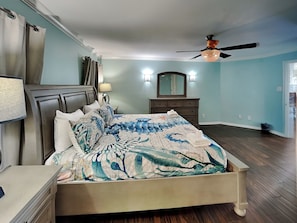 A bedroom with a bed, a dresser, a mirror, a ceiling fan, and a window. - Harbor View Breeze by Ryson Vacation Rentals (Galveston)