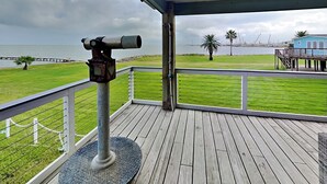 A wooden deck with a telescope, a grassy area, and a view of the water and distant structures. - Harbor View Breeze by Ryson Vacation Rentals (Galveston)