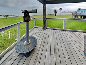 A wooden deck with a telescope, a grassy area, and a view of the water and distant structures. - Harbor View Breeze by Ryson Vacation Rentals (Galveston)