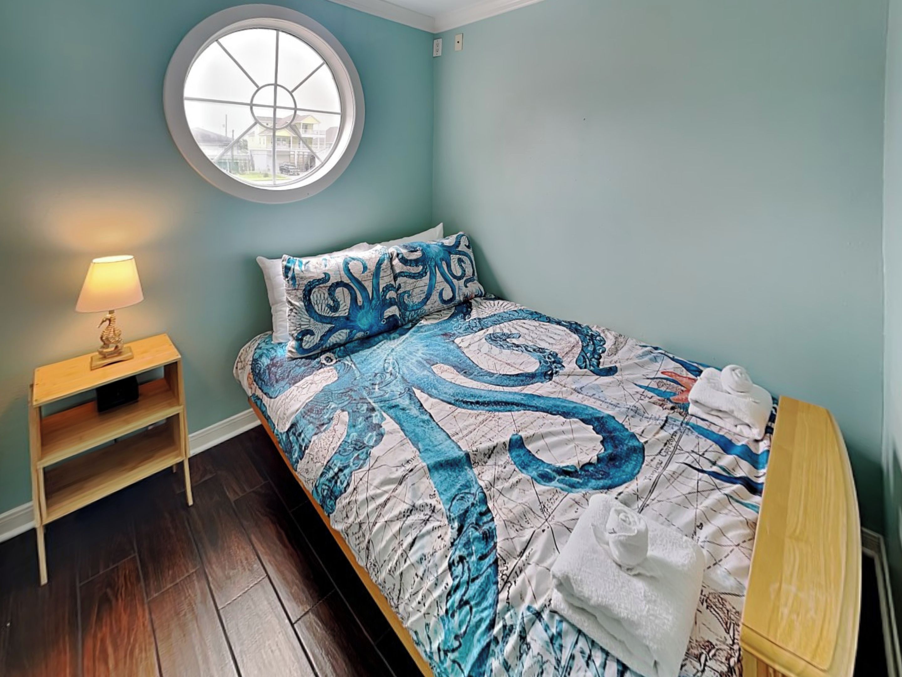 A bedroom with a bed, a nightstand, a lamp, a window, and a view of houses outside.