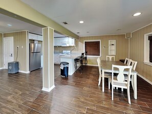 A spacious living area with a dining table and chairs, a kitchen with modern appliances, and a large window with blinds. - Harbor View Breeze by Ryson Vacation Rentals (Galveston)