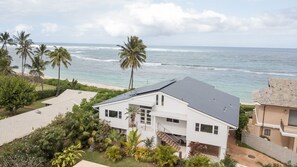 Ein modernes, zweistöckiges Haus mit geneigtem Dach, großen Fenstern und einem Balkon mit Blick auf einen Strand mit Palmen und Wellen.