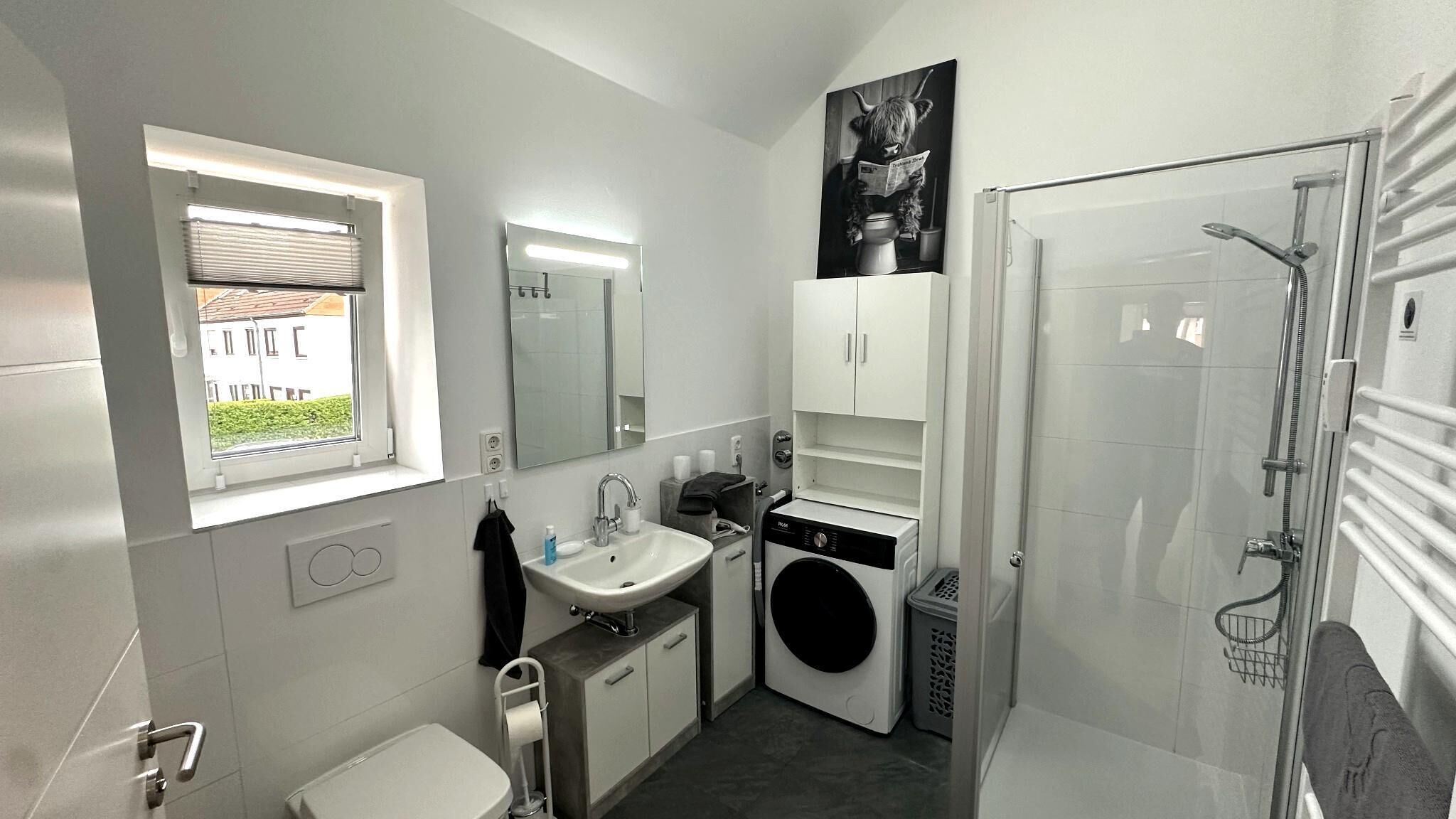 A modern bathroom with a window, a sink, a mirror, a toilet, a washing machine, and a shower.