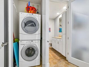 A laundry area with two stacked washing machines, a sink, and a mirror in the adjacent room. - The Cozy Corner by Ryson Vacation Rentals (Galveston)