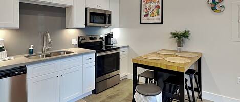 A modern kitchen with white cabinets, stainless steel appliances, a wooden dining table, and a trash can.