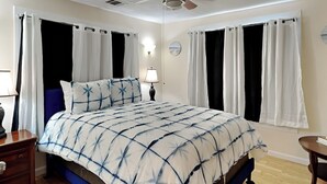 A bedroom with a bed, curtains, a nightstand, a lamp, and a small table. - Sugar Shack Bungalow by Ryson Vacation Rentals (Galveston)