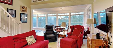 A spacious living room with a red sofa, a black leather chair, and a wooden coffee table. There is a dining area with a table and chairs, and a TV mounted on the wall.