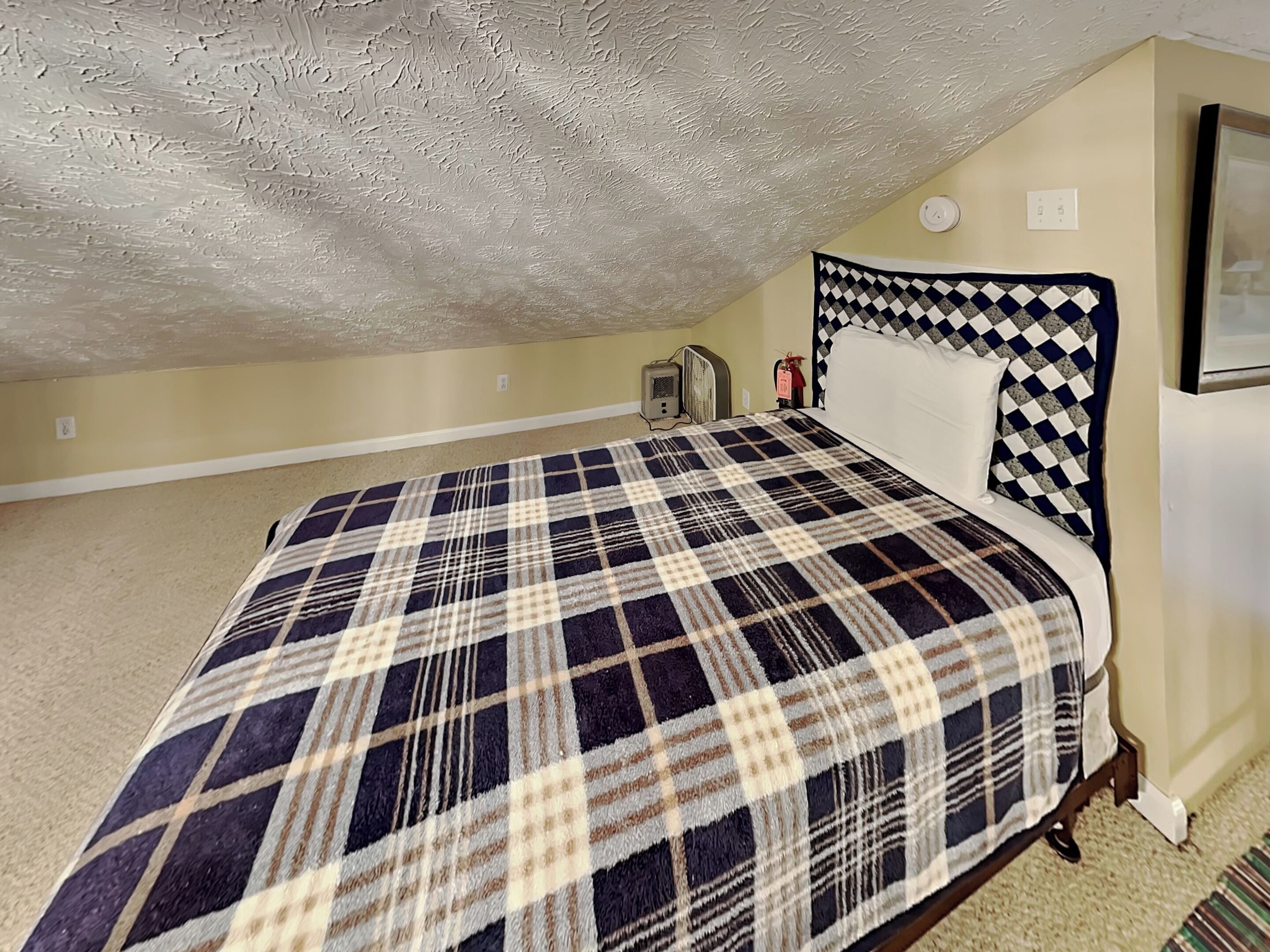 A bedroom with a checkered bedspread, a headboard with a diamond pattern, a small heater, and a wall-mounted light.
