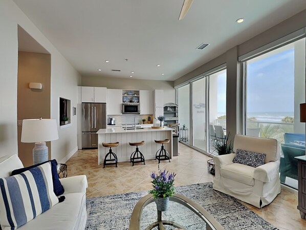 A modern living room with a sofa, a coffee table, and a view of the kitchen and balcony. - Endless Summer by Ryson Vacation Rentals (Galveston)