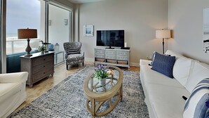 Un salon spacieux avec une grande fenêtre, un ventilateur de plafond, un canapé blanc, une table basse ornée d’un vase et un téléviseur posé sur un meuble.
