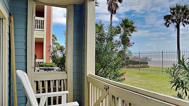 A porch with white Adirondack chairs, a view of a beach, and a fence. - First Light by Ryson Vacation Rentals (Galveston)