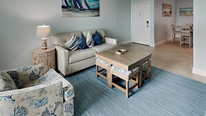 A living room with a sofa, a coffee table, and a patterned armchair. There is a painting on the wall and a dining area in the background. - First Light by Ryson Vacation Rentals (Galveston)