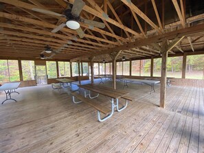 A covered picnic area with wooden benches and tables, a ceiling fan, and large windows offering views of the surrounding trees.