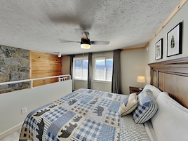 Un dormitorio con cama, ventilador de techo, ventana con cortinas y pared de piedra.