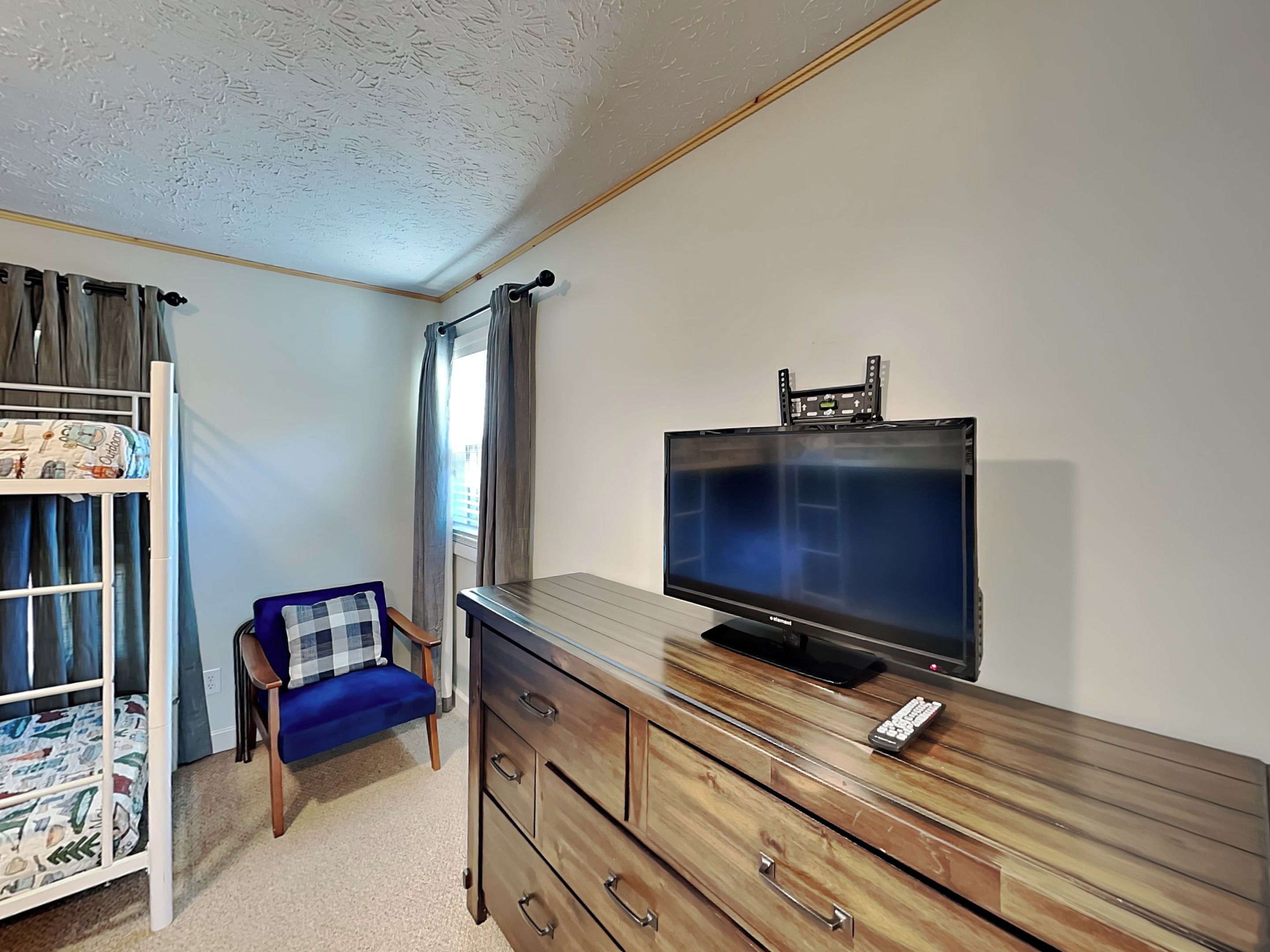 Een slaapkamer met een stapelbed, een tv op een houten dressoir, een stoel en een raam met gordijnen.