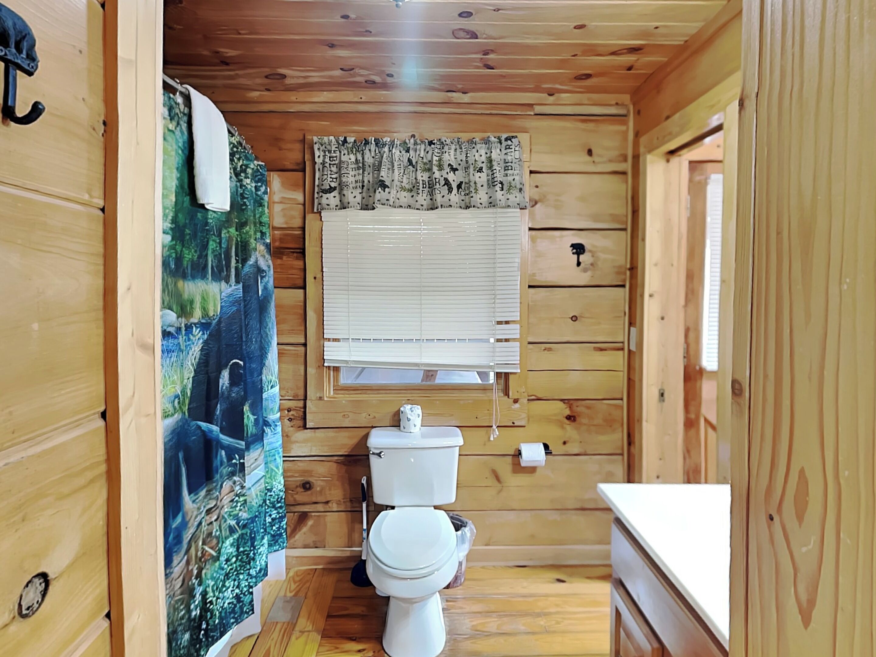 A wooden cabin bathroom with a toilet, a window with blinds, a shower curtain with a forest scene, and a towel hanging on the wall.